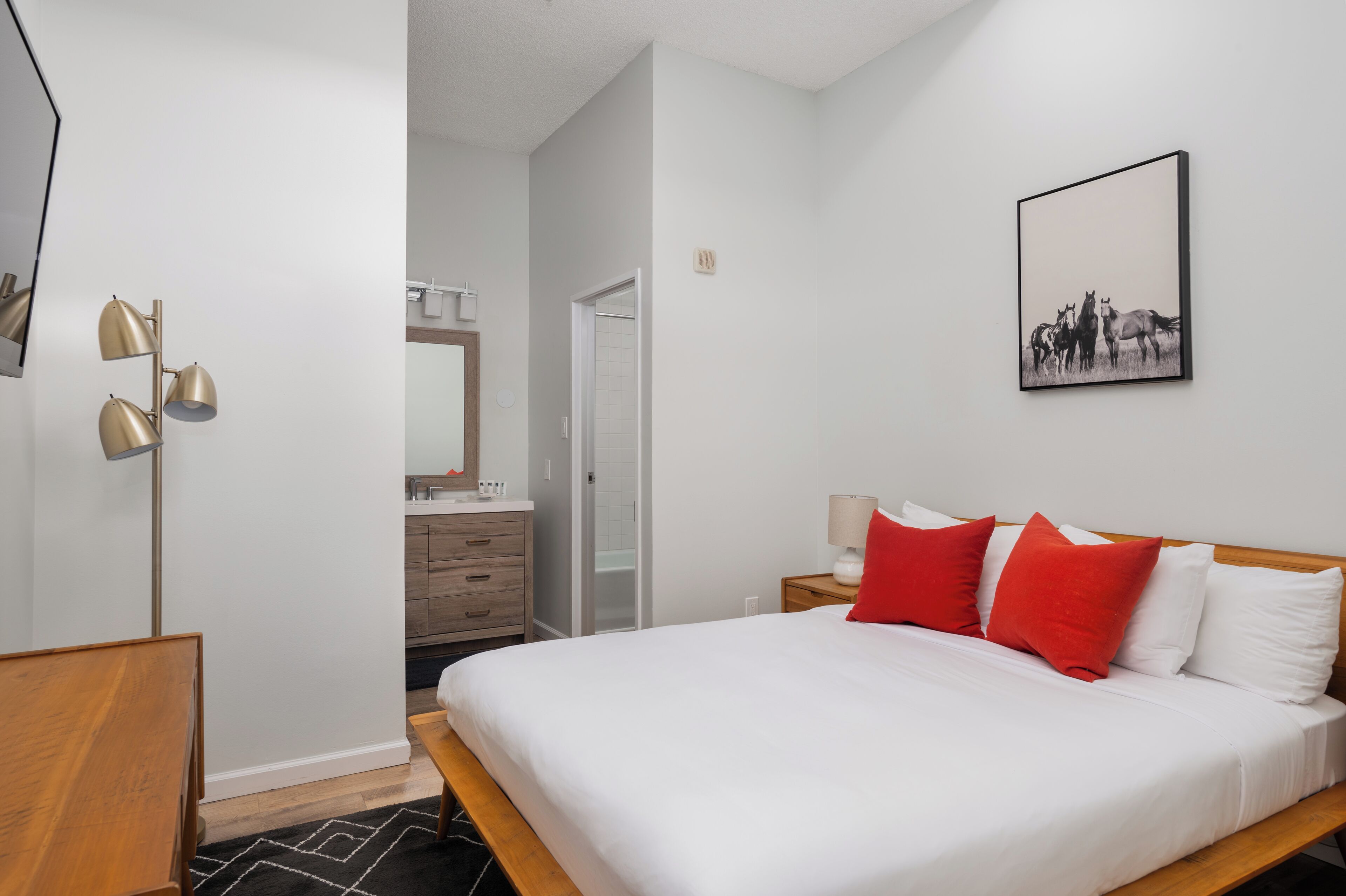 A hotel room with a bed, a bedside table, a lamp, a mirror, a bathroom, and a hallway.