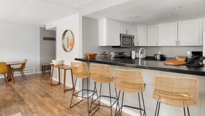 Cocina moderna con armarios blancos, encimera negra y microondas de acero inoxidable. Hay tres taburetes de mimbre y una mesa de madera con sillas.