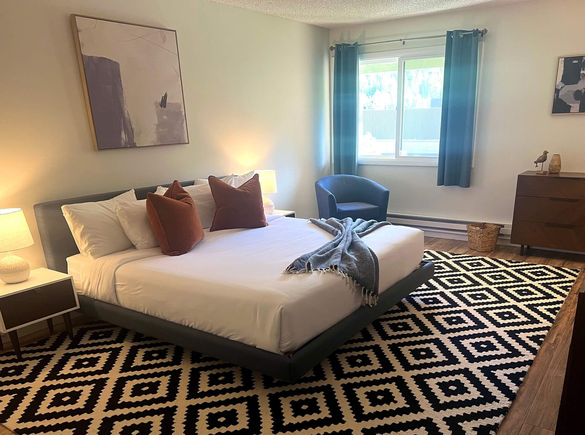 A hotel room with a bed, a chair, a dresser, and a window with curtains.