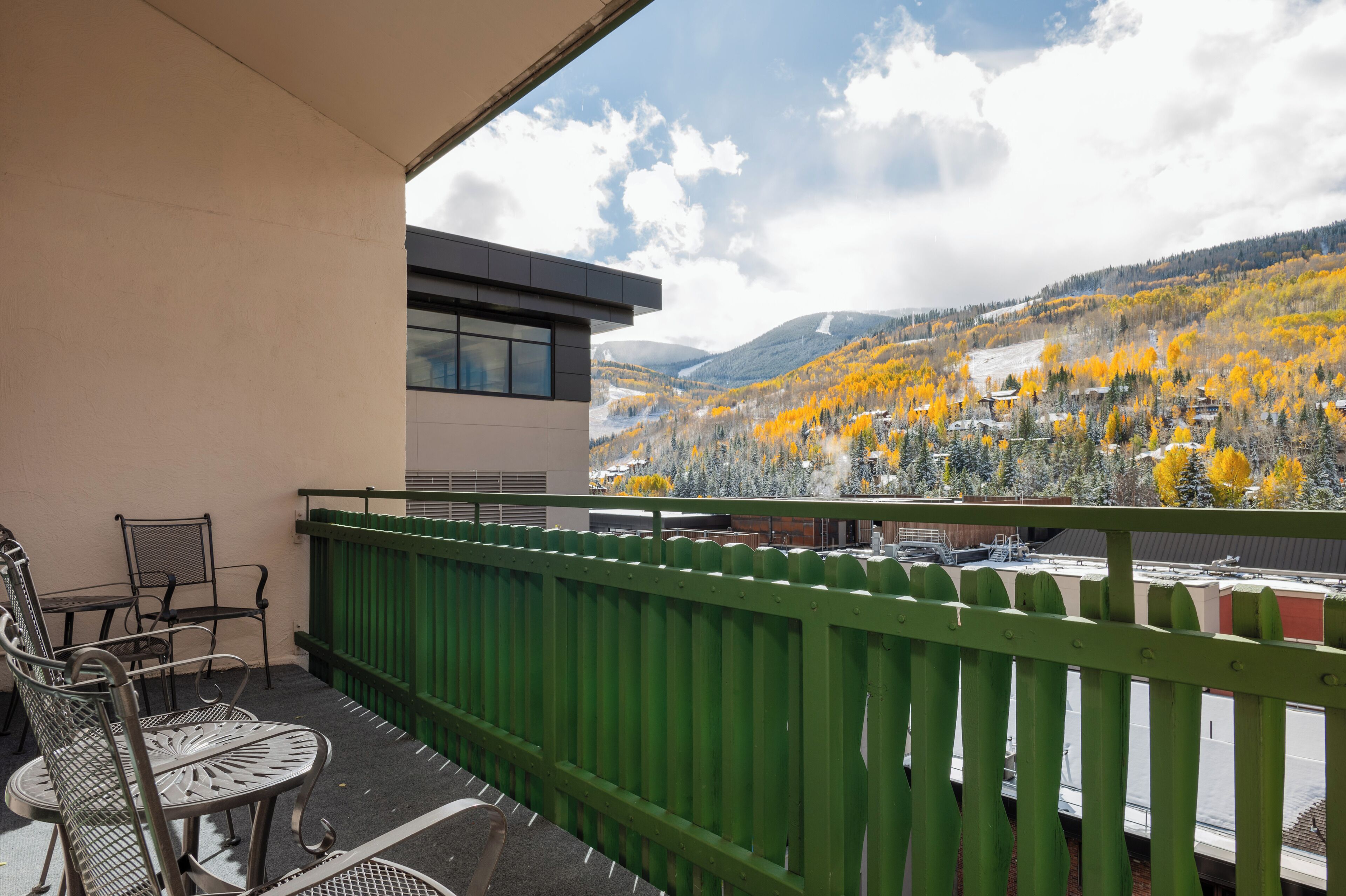 A balcony with a green railing, metal chairs, and a view of a mountainous landscape with autumn foliage.