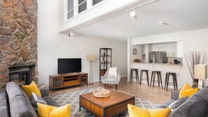 Una sala de estar con chimenea de piedra, televisor, zona de bar y cocina al fondo.