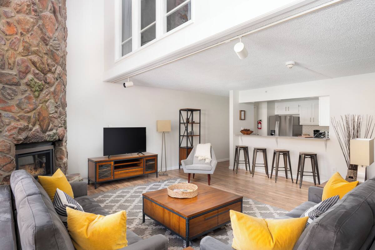 Una sala de estar con chimenea de piedra, televisor, zona de bar y cocina al fondo.