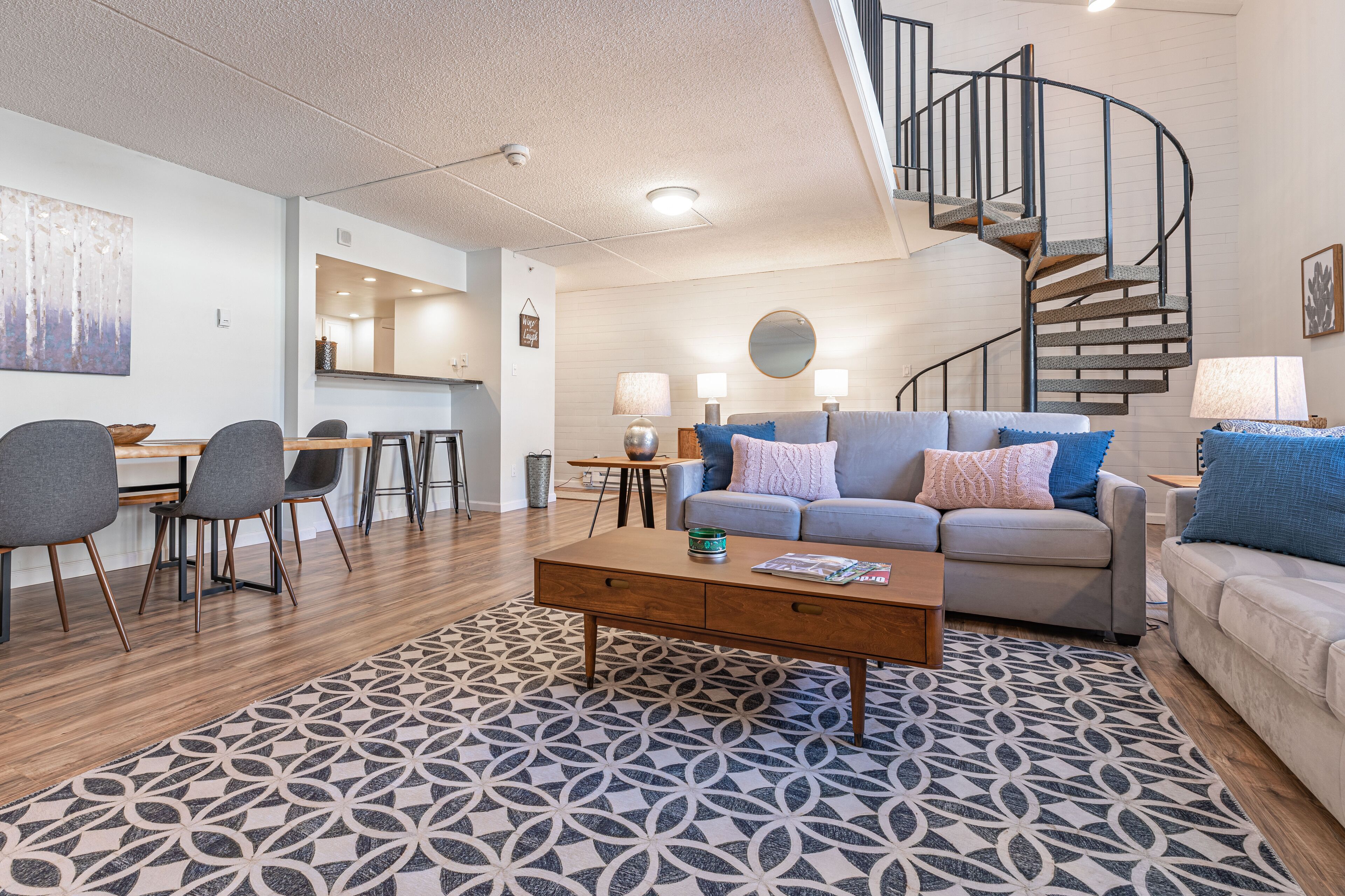 A modern living room with a spiral staircase, a dining area, and a large abstract painting on the wall.