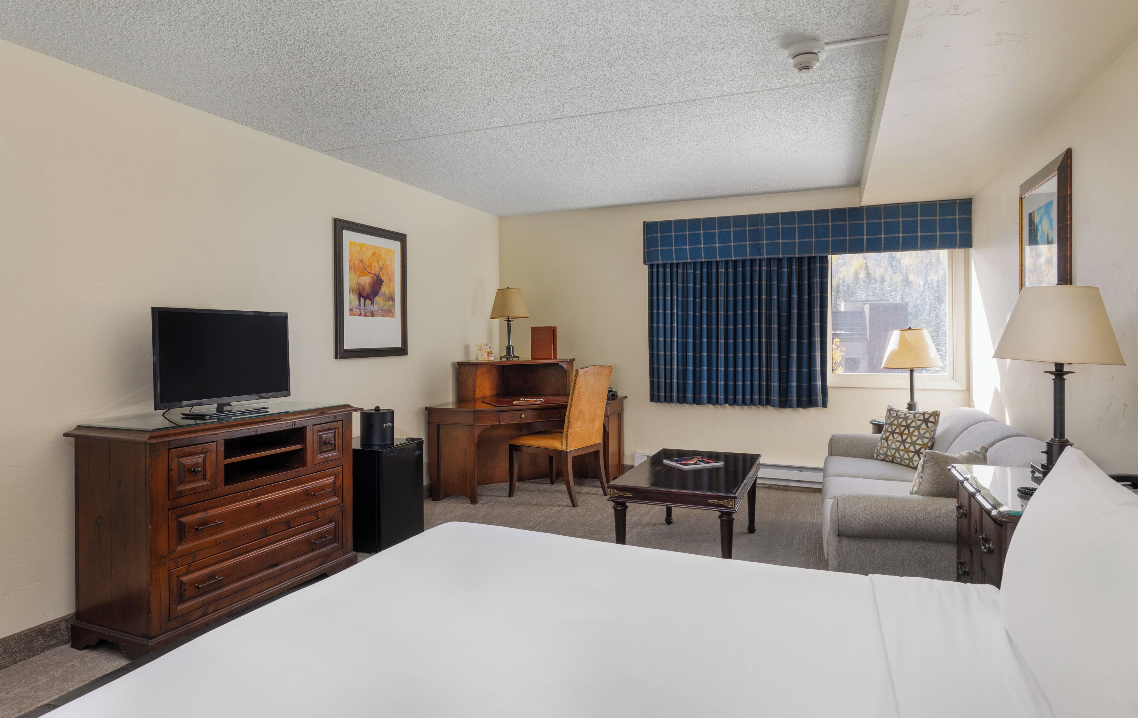 Una habitación de hotel con cama, escritorio, silla, televisor, lámpara y ventana con cortinas.