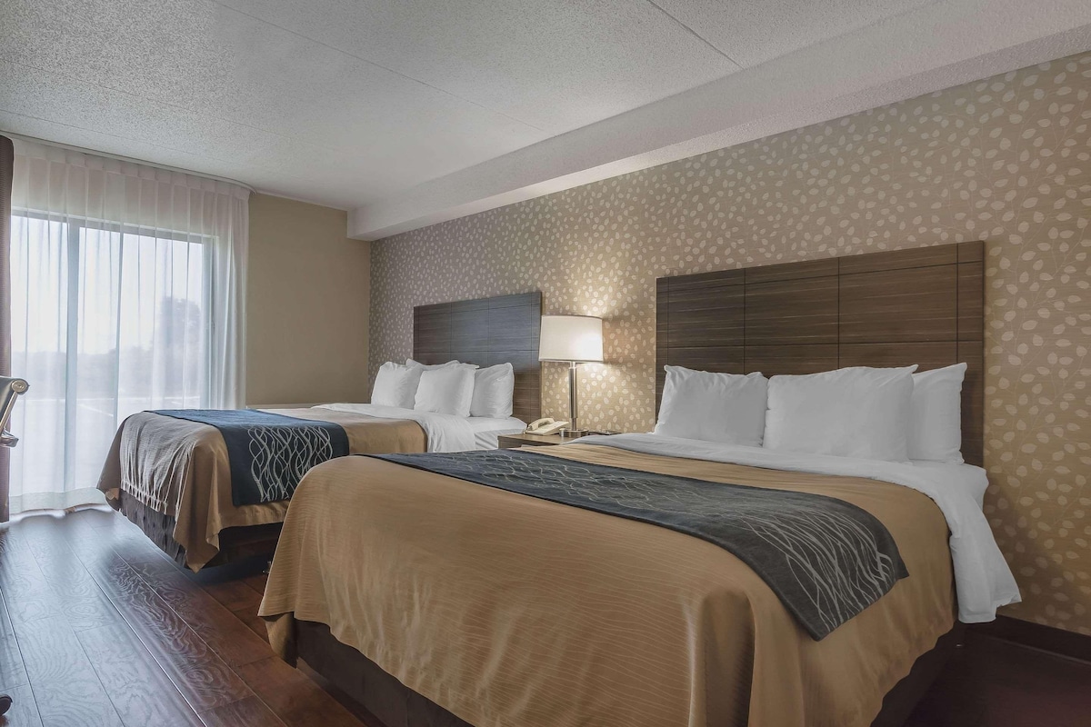 A hotel room with two beds, a nightstand, a lamp, and a window with curtains.