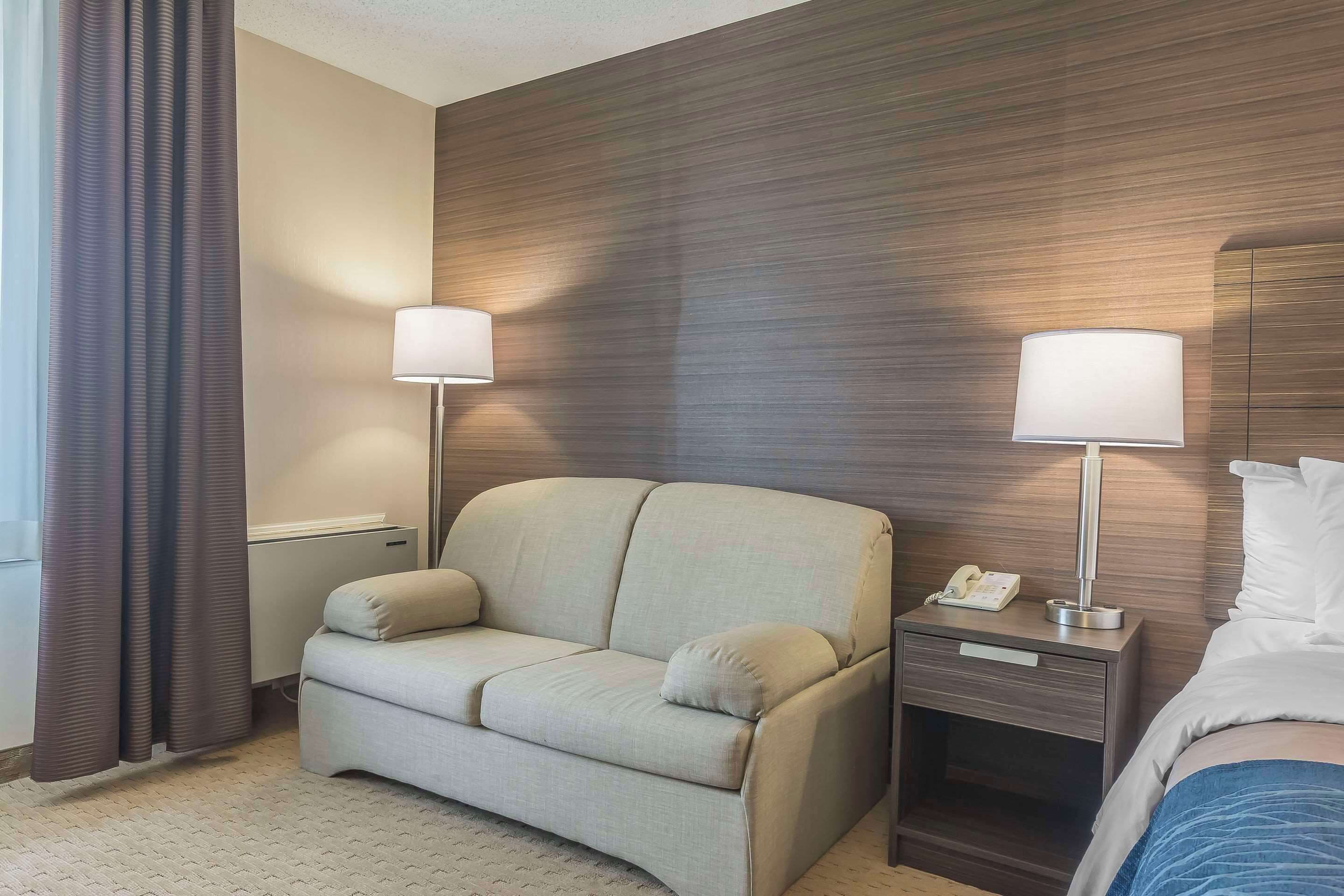 A hotel room with a sofa, a bedside table with a lamp, a phone, and a bed.
