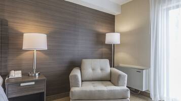 Una habitación de hotel con un sillón gris, una mesita de noche con lámpara y un teléfono.