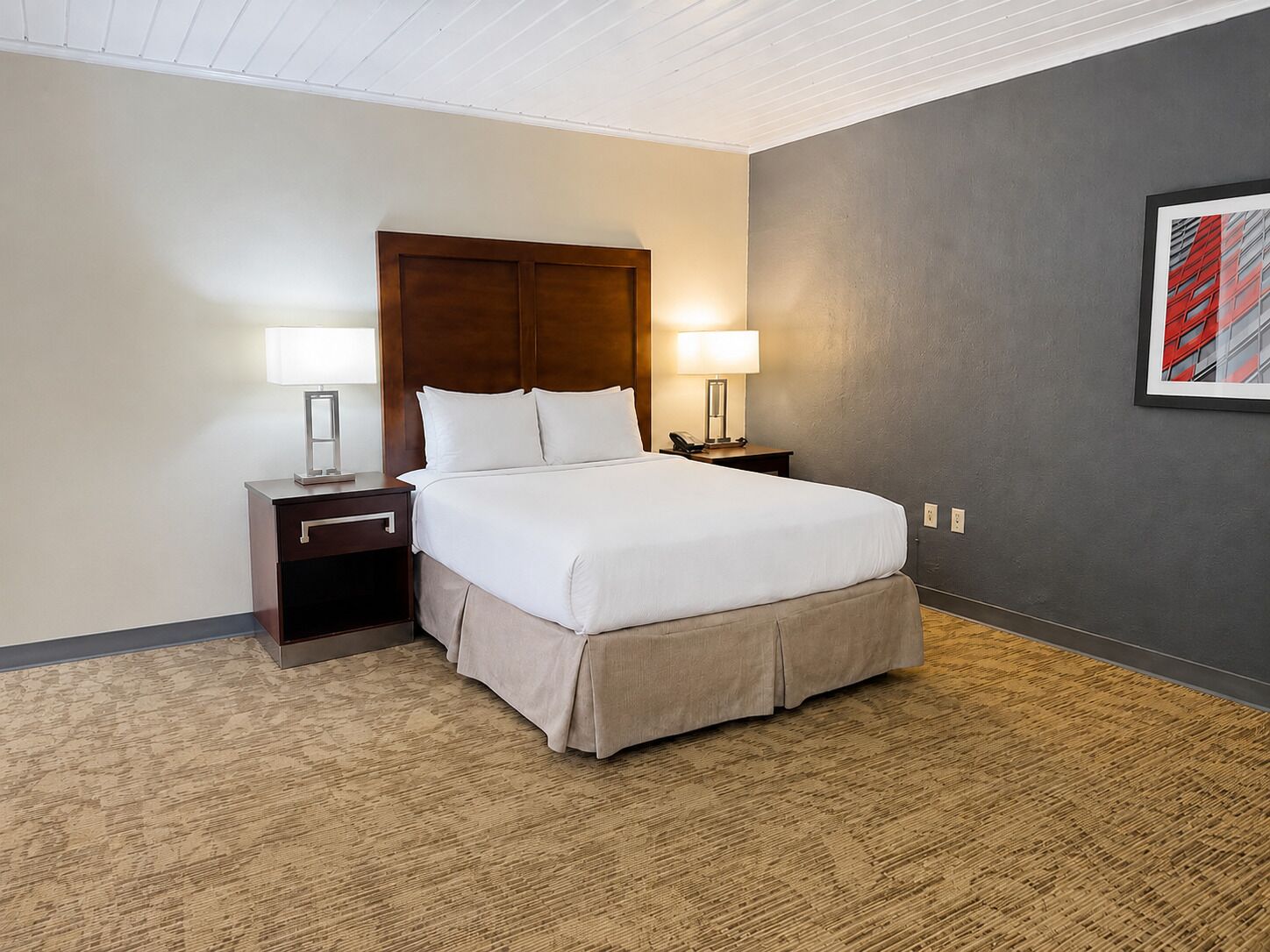 Quarto de hotel com uma cama grande, duas mesas de cabeceira com abajures, uma parede escura e um quadro emoldurado.