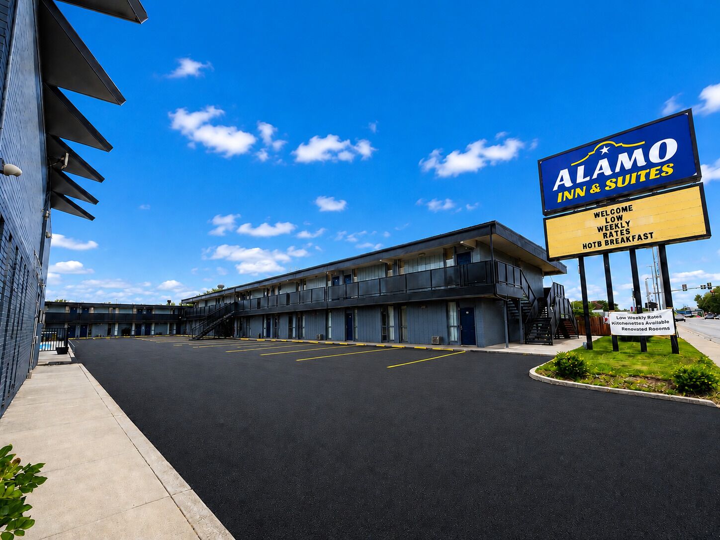 Un motel avec une enseigne publicitaire indiquant « Alamo Inn & Suites », avec un bâtiment moderne doté d’un toit plat, un parking et un ciel bleu dégagé.