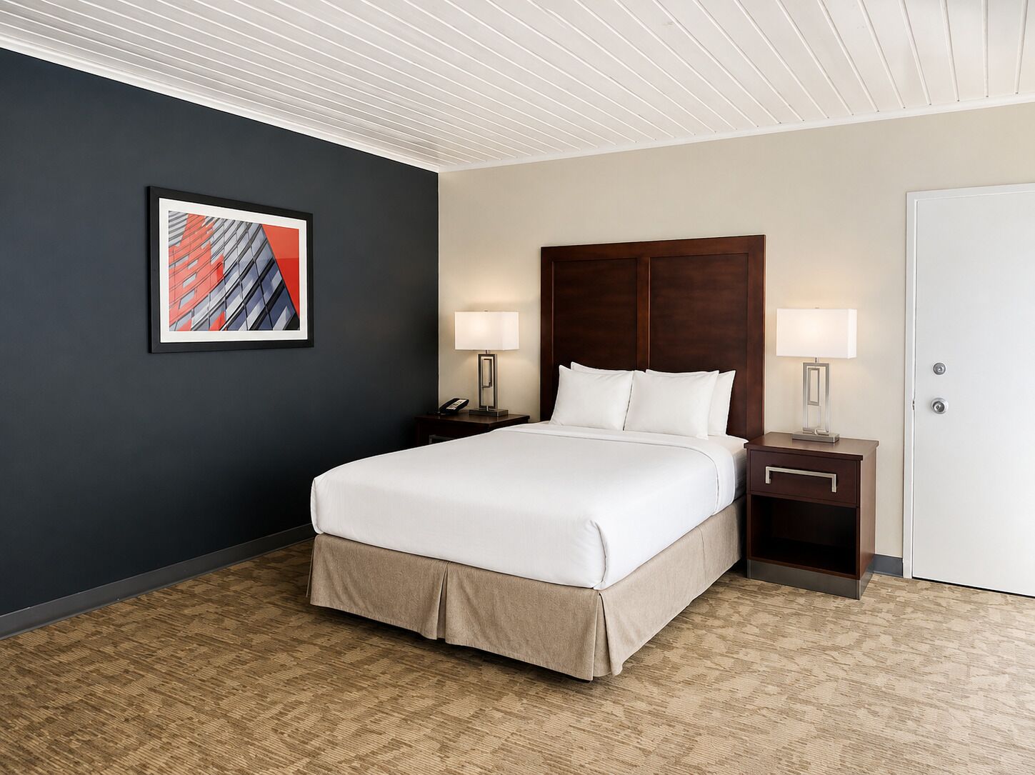 Una habitación de hotel con una cama, dos mesitas de noche con lámparas, una mesita auxiliar y un cuadro en la pared, además de una puerta.