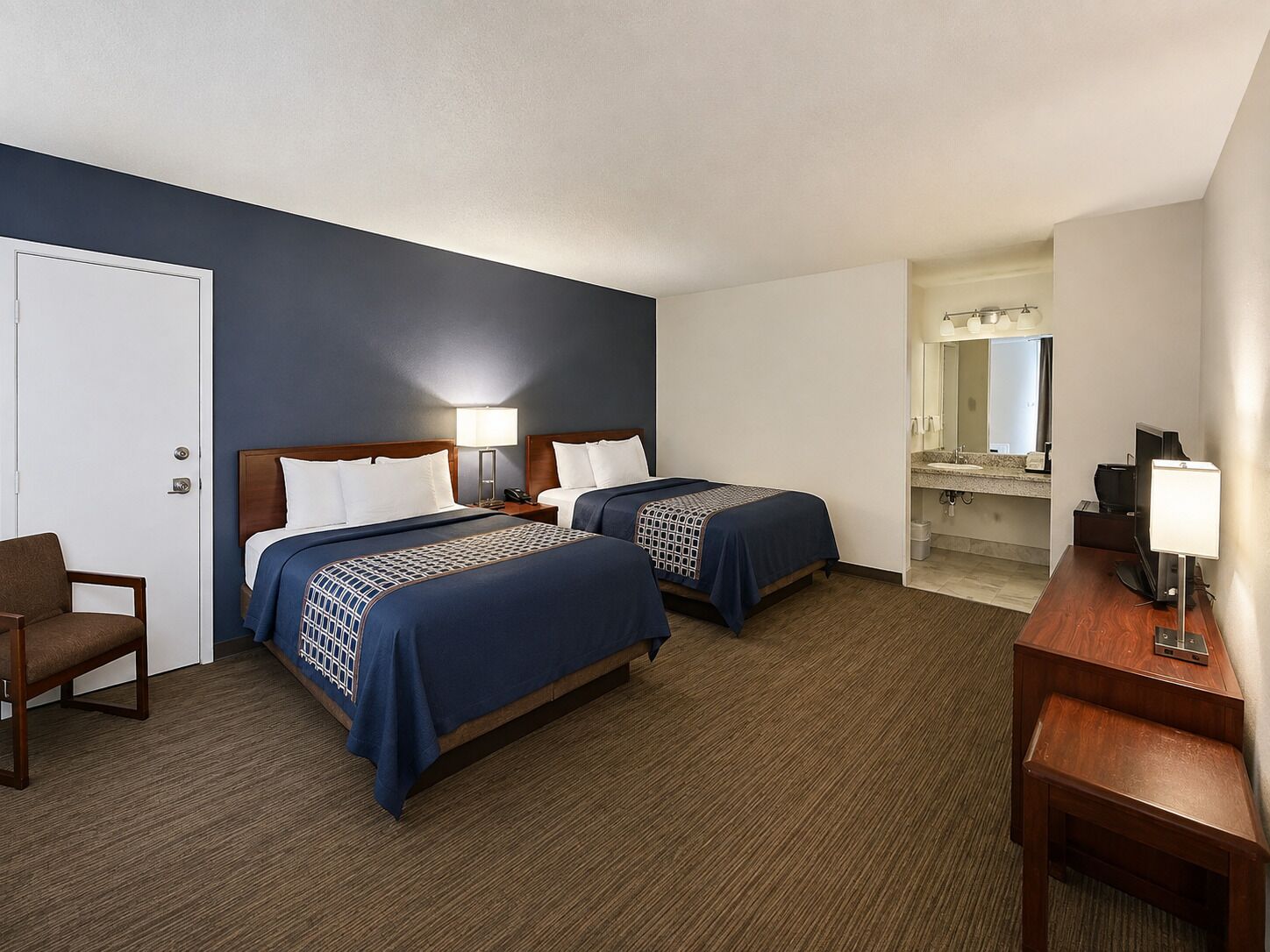 A hotel room with two beds, a desk, a chair, a bathroom with a mirror, and a TV.