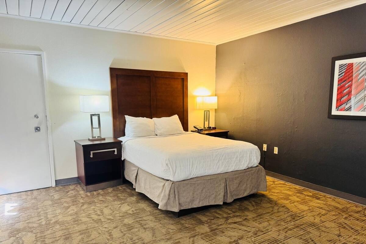 A hotel room with a bed, two bedside tables, a lamp, a framed picture on the wall, and a door.