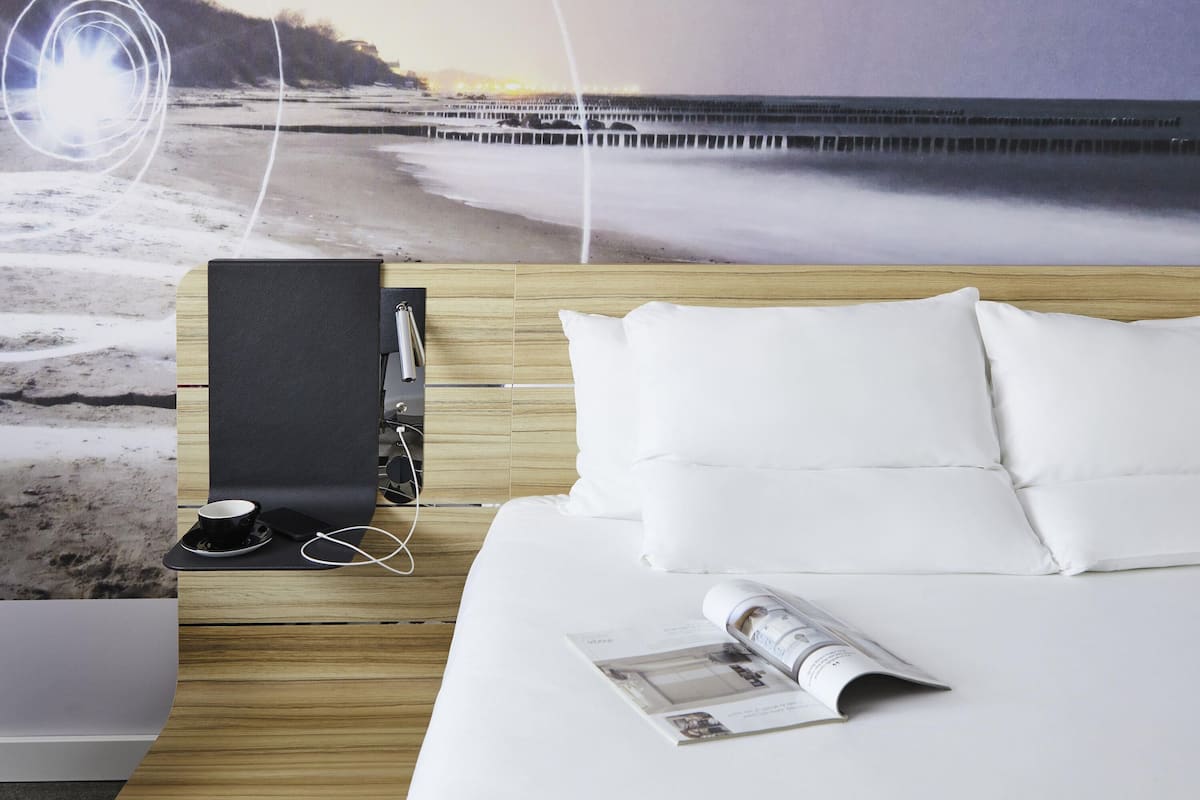 Een slaapkamer met een houten hoofdbord, wit beddengoed, een tijdschrift op het bed, een zwart kussen op het nachtkastje en een koffieapparaat.