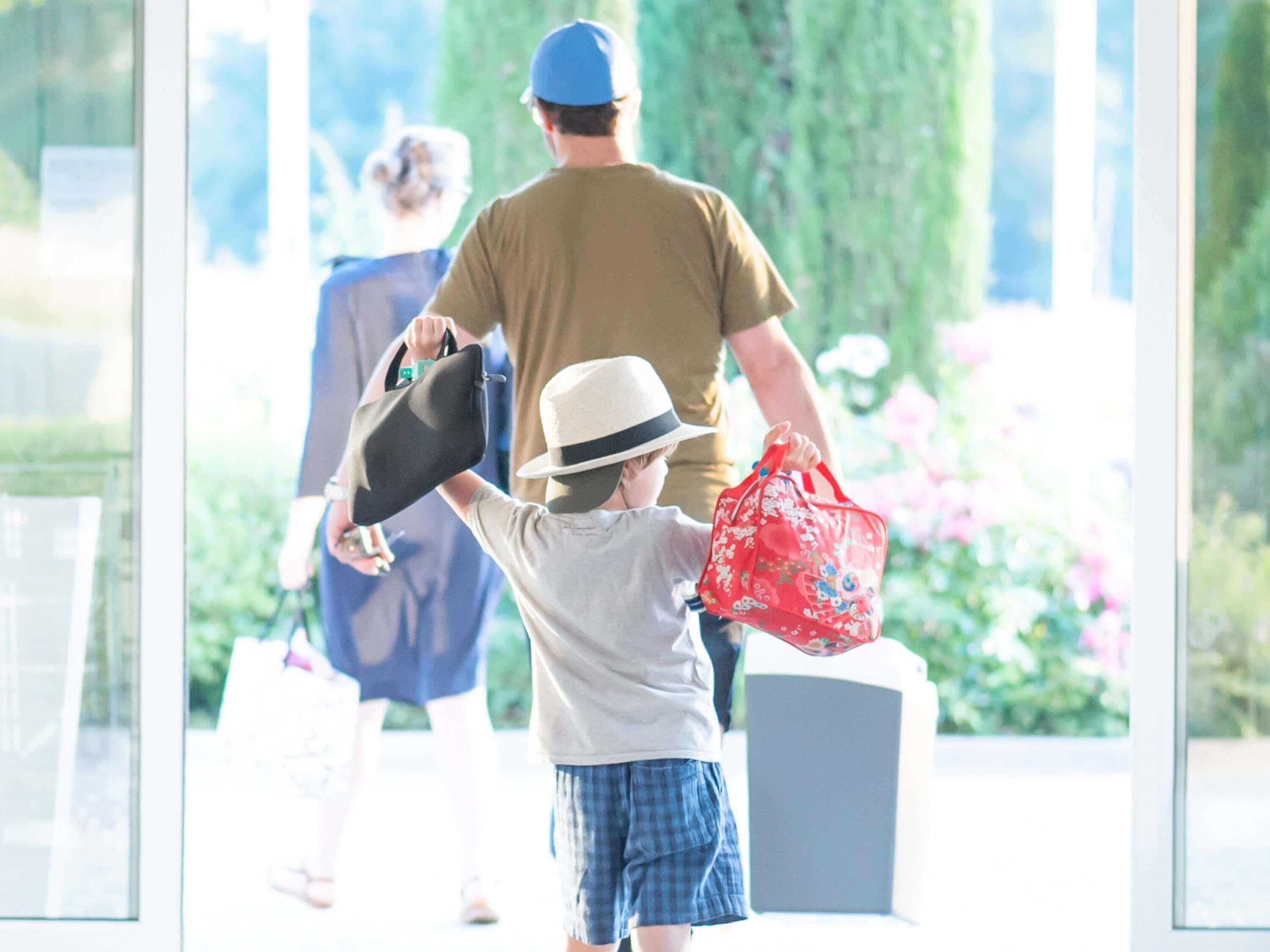 A child holding a hat and a red bag, with an adult in the background.