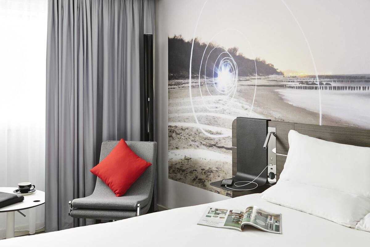 A hotel room with a bed, a reading area with a magazine, a chair with a red pillow, and a large wall mural of a beach scene.