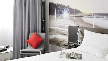 A hotel room with a bed, a reading area with a magazine, a chair with a red pillow, and a large wall mural of a beach scene.