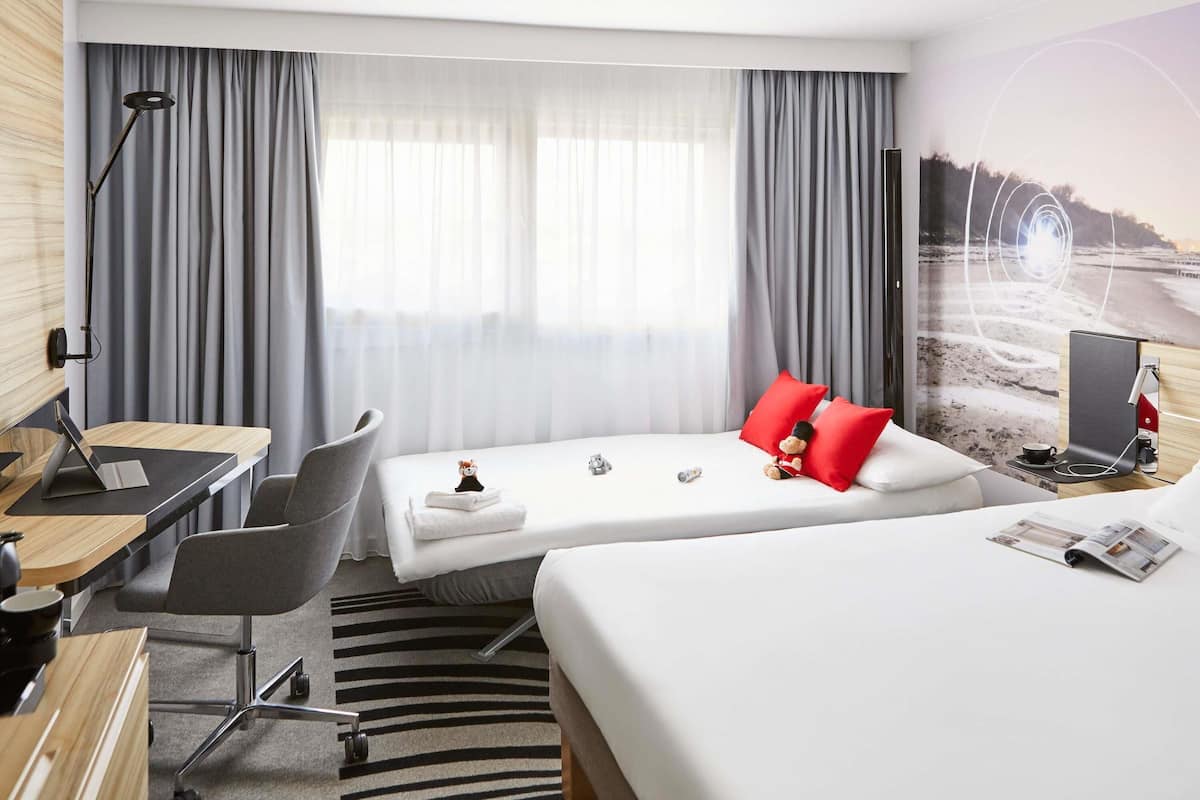 Hotelkamer met een bureau, twee bedden, een stoel, een lamp en een groot raam met gordijnen.
