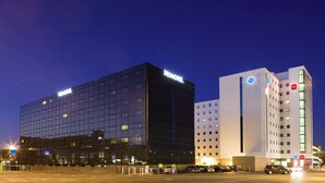 Ein modernes Hotel mit Glasfassade, Parkplatz und angrenzenden Gebäuden mit sichtbarer Beschilderung.