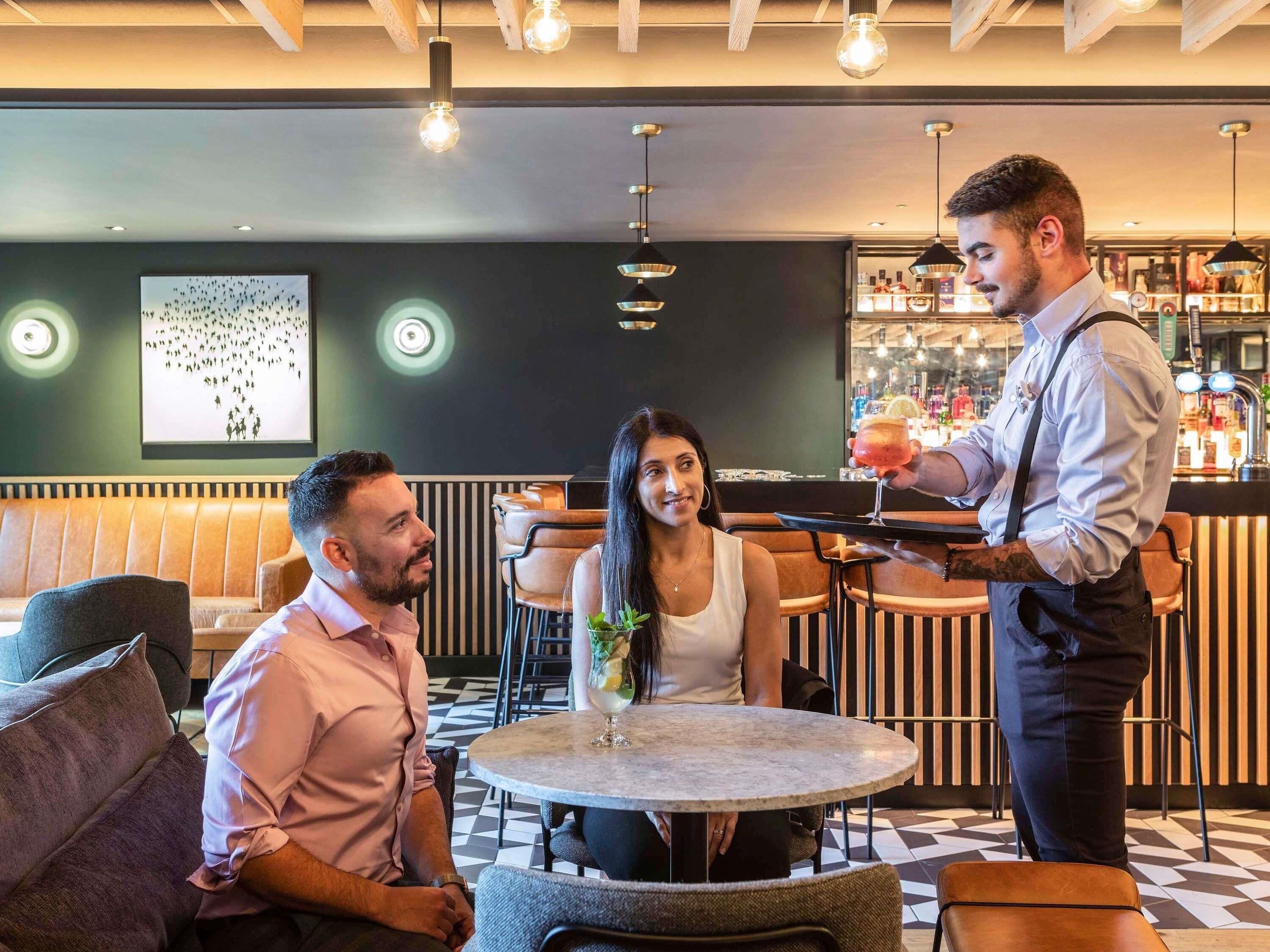 Uma área de bar com uma mesa redonda, banquetas e um barman servindo bebidas.
