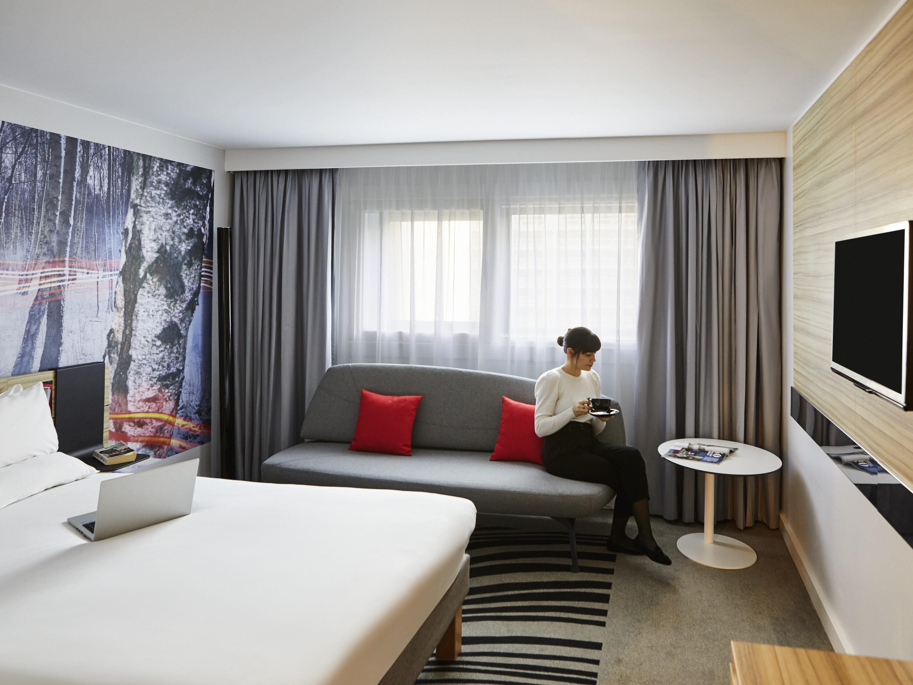 Ein modernes Hotelzimmer mit einer Couch, einem Bett, einem Fernseher und einem Laptop auf dem Bett.