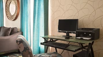 A room with a glass desk, a computer, and a printer. There is a chair with a patterned cushion and a sofa. The room has a patterned wallpaper and curtains.