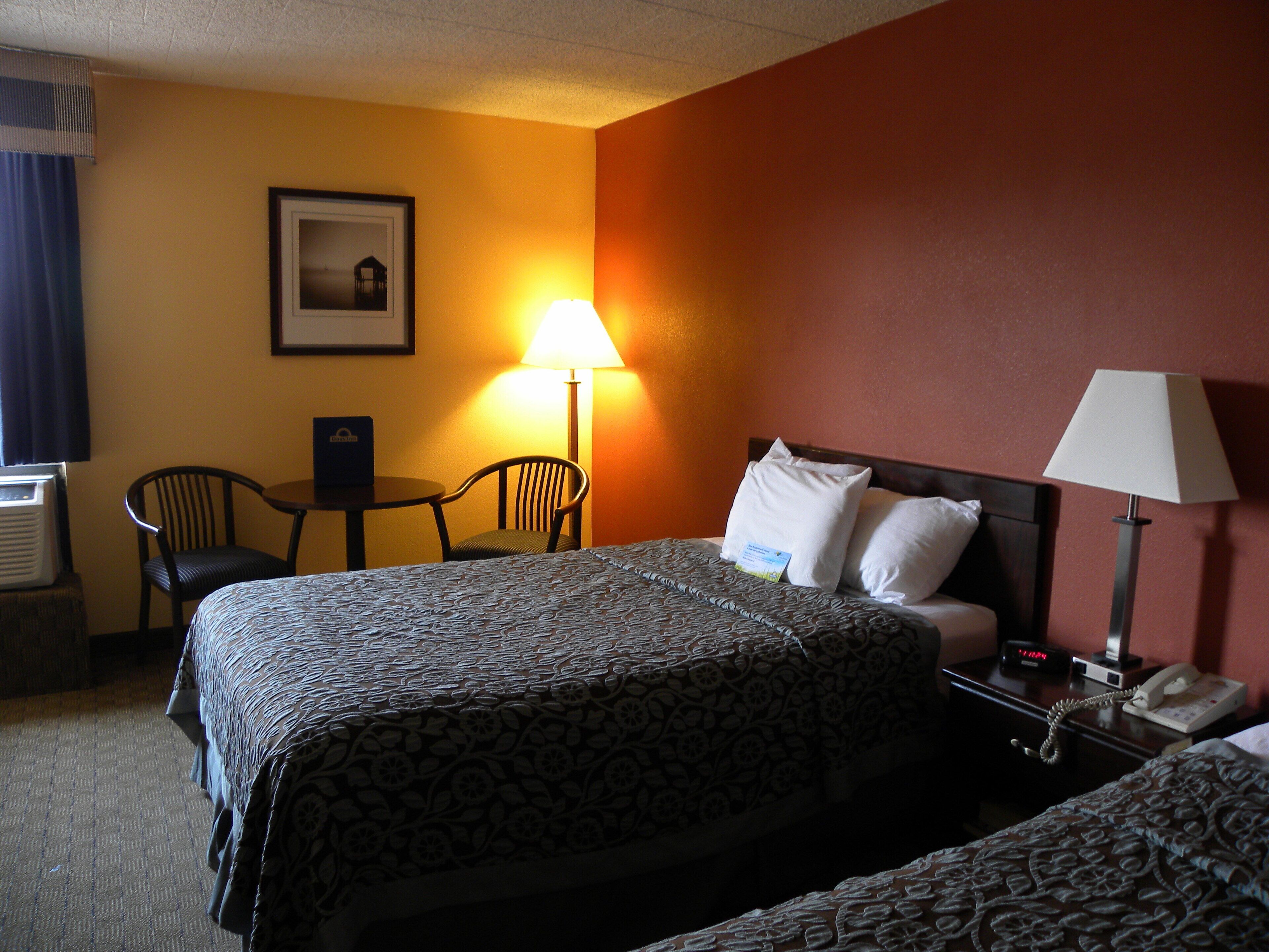 A hotel room with two beds, a desk, a chair, a lamp, and a framed picture on the wall.