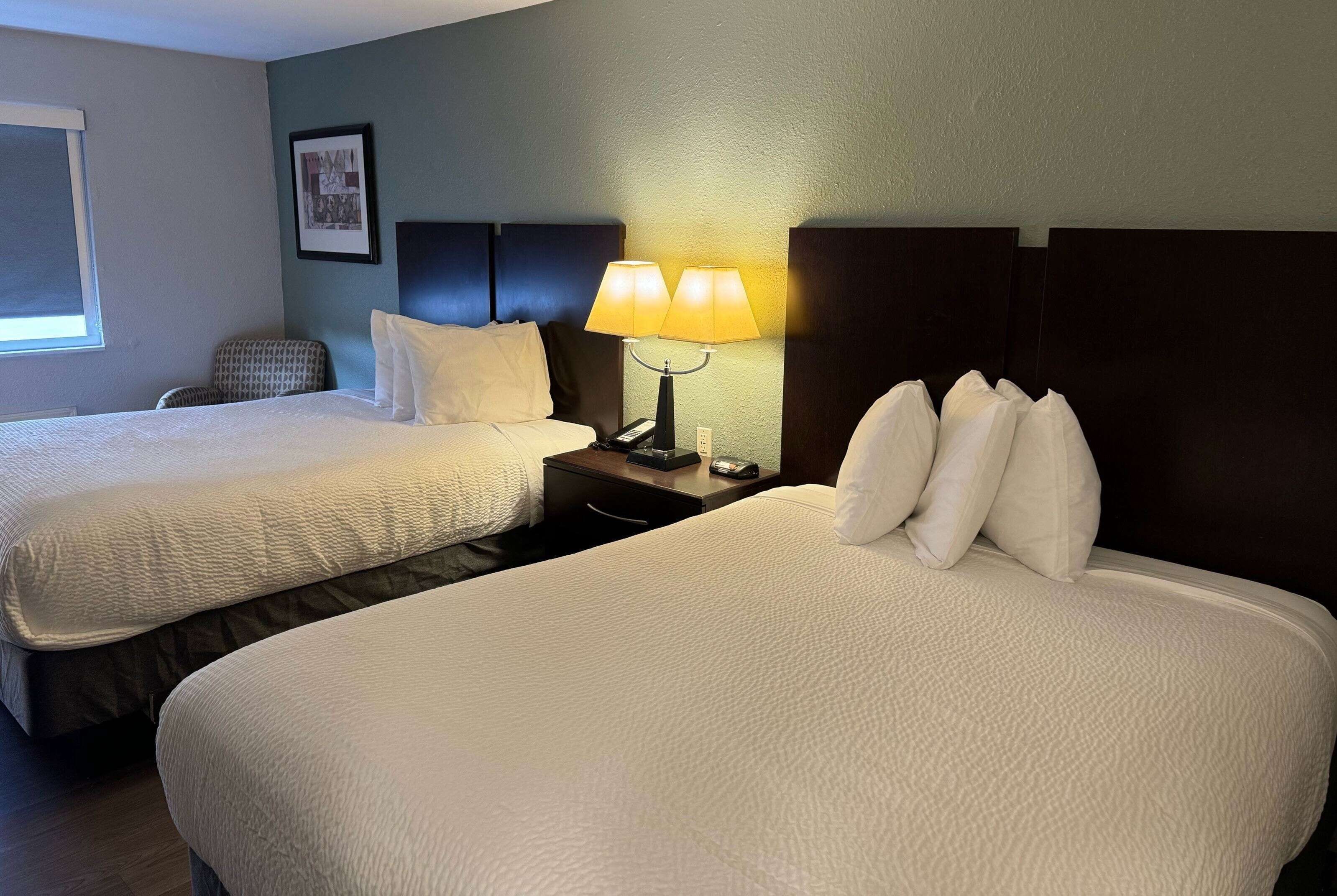 A hotel room with two beds, a nightstand with a lamp, a framed picture on the wall, and a window with blinds.