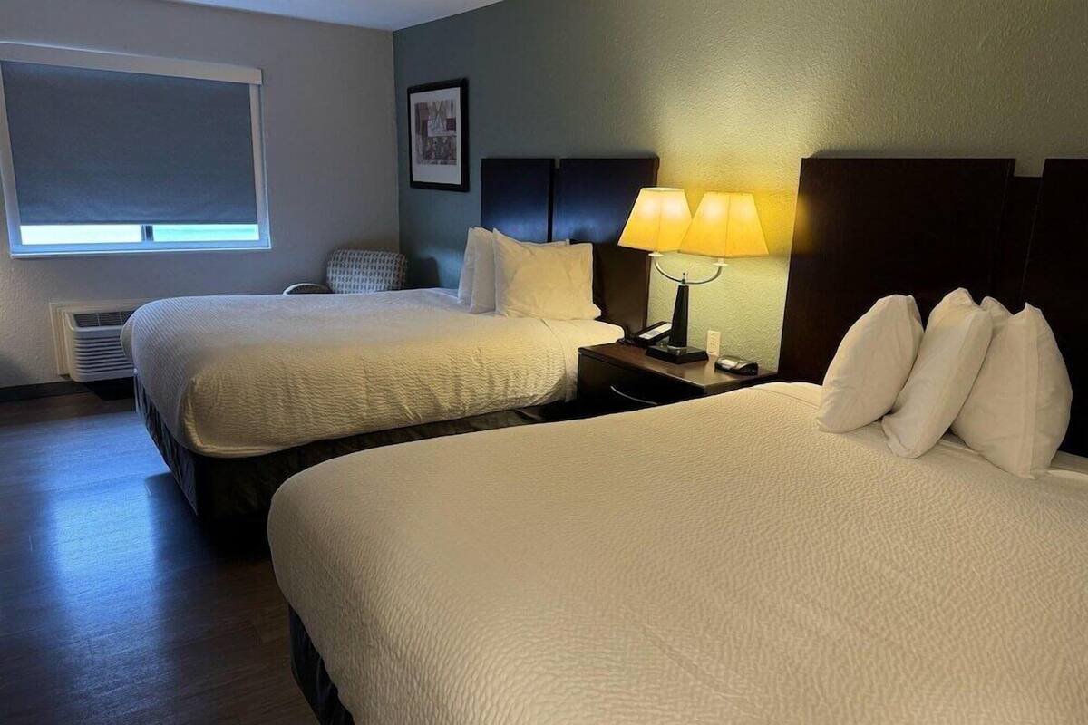 A hotel room with two beds, a bedside table with lamps, a chair, and a window with blinds.