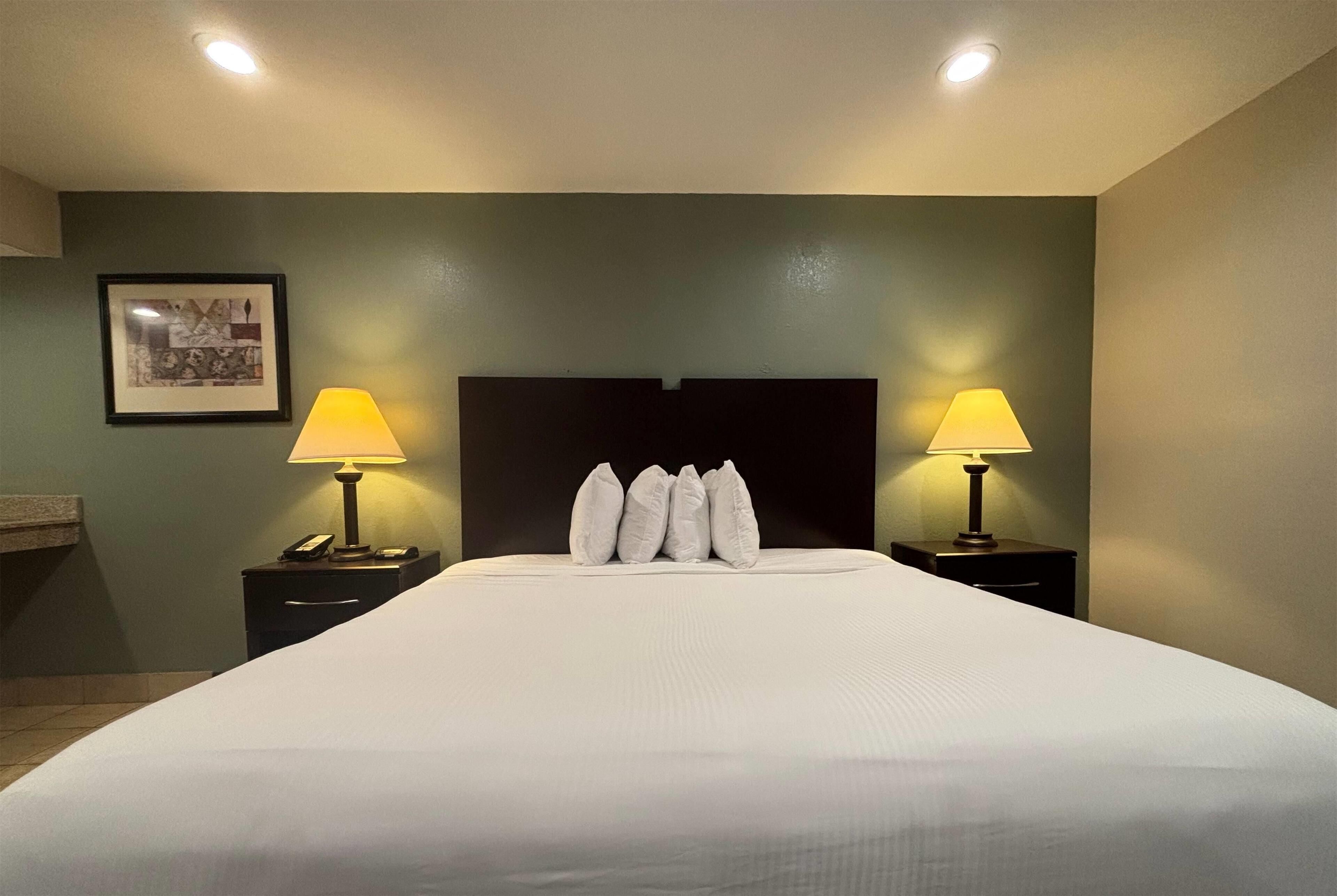 A hotel room with a large bed, two bedside tables with lamps, a framed picture on the wall, and a dark headboard.
