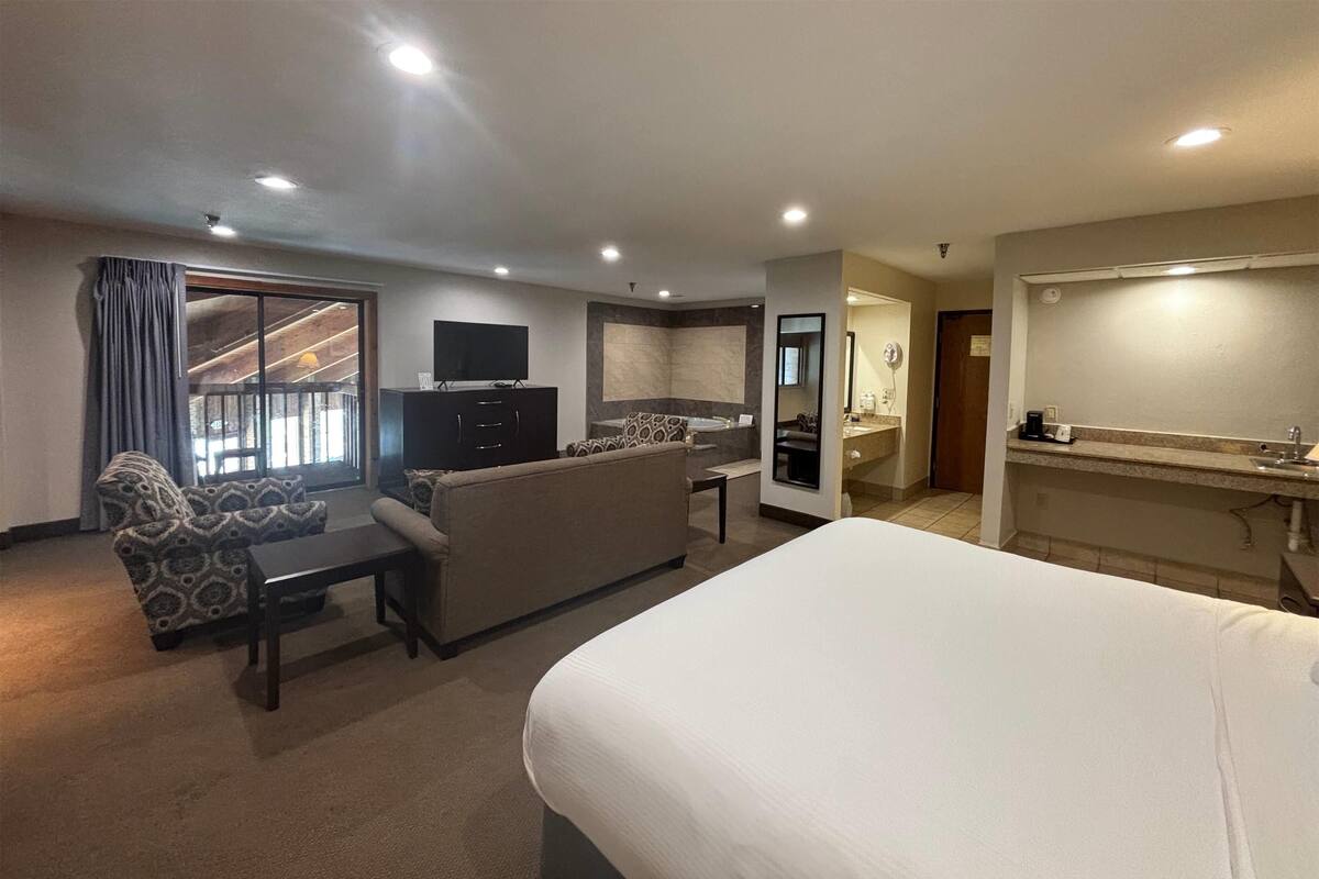 A hotel room with a large bed, a sofa, a desk, a television, and a kitchen area.