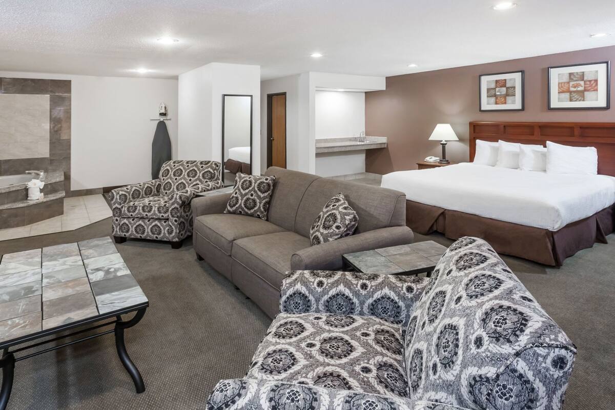 A hotel room with a bed, two sofas, a coffee table, a bathroom with a shower, and a mirror.