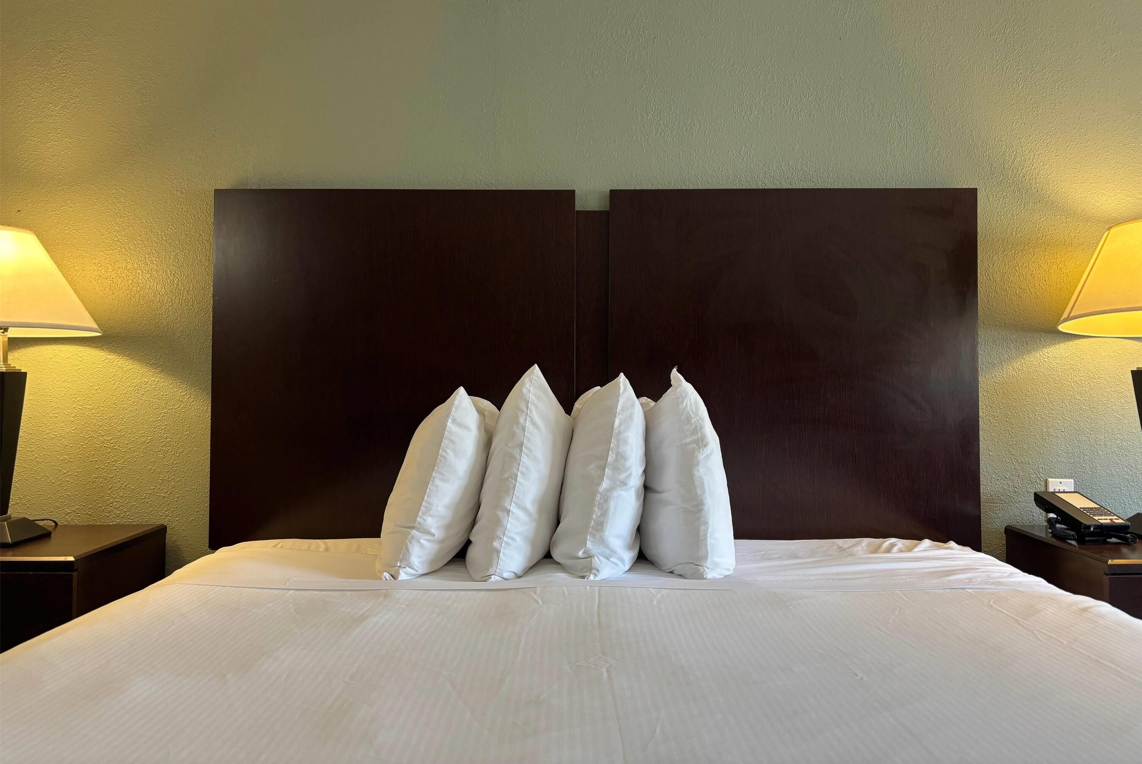 A neatly made bed with white linens and a dark wooden headboard. There are four white pillows aligned at the head of the bed. Two bedside tables flank the bed, each with a lamp and a phone.