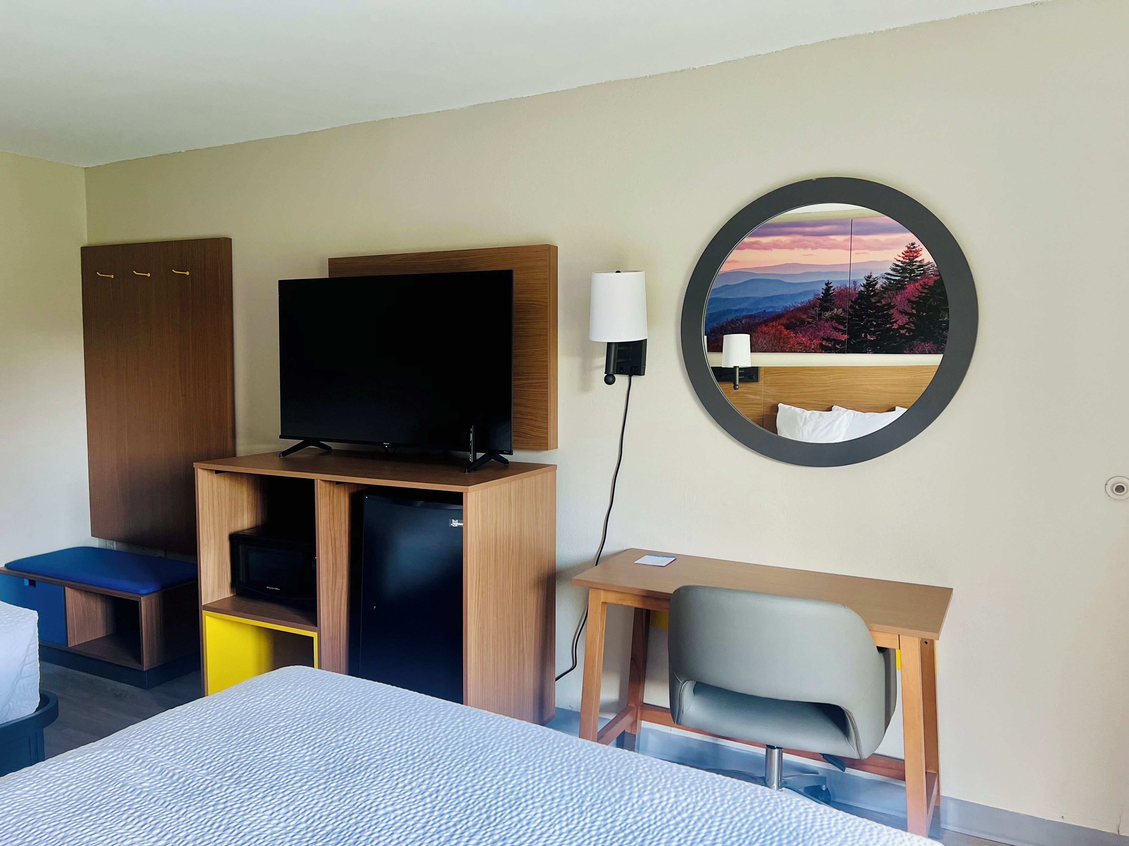 A hotel room with a bed, a desk with a chair, a television, a wardrobe, and a round mirror with a scenic view.
