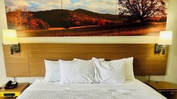 Een hotelkamer met een houten hoofdeinde, wit beddengoed, een landschapsschilderij boven het bed en bedlampen.