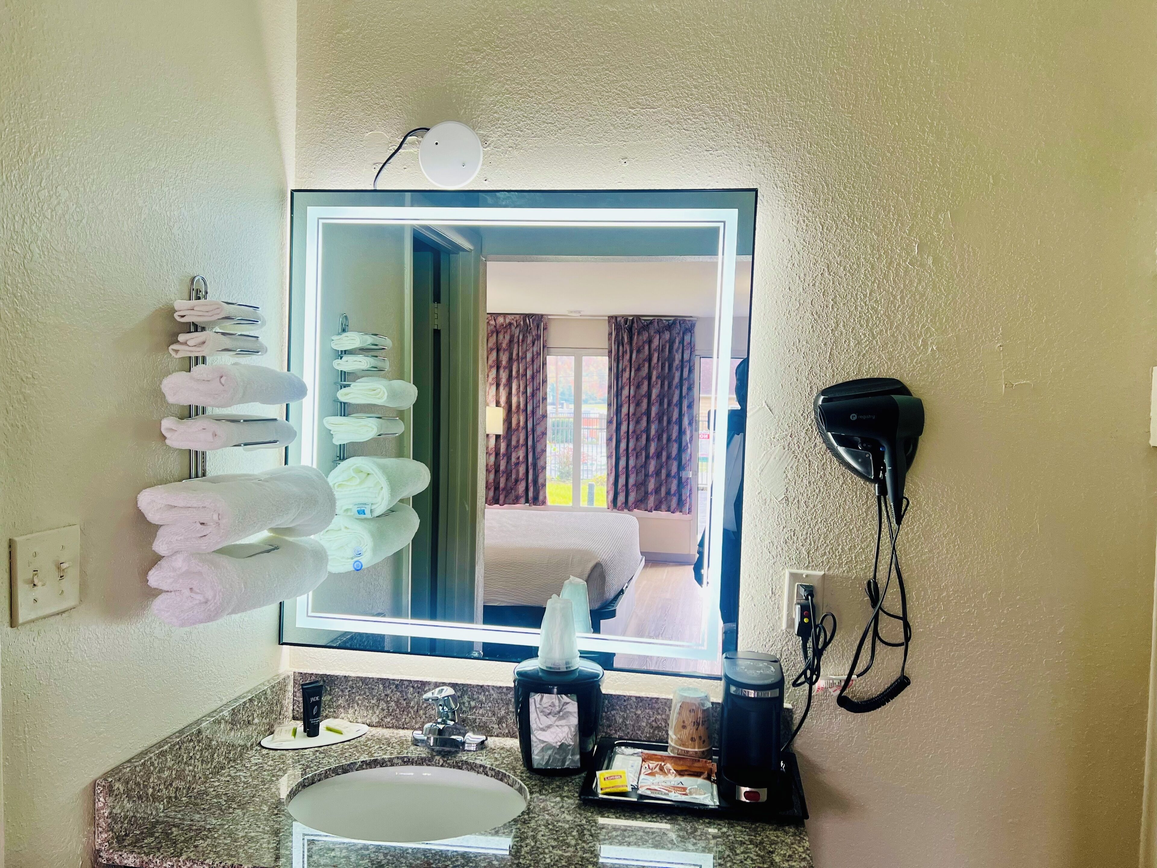A hotel bathroom with a large mirror, a towel rack with towels, a hairdryer, a sink with toiletries, and a window with curtains.