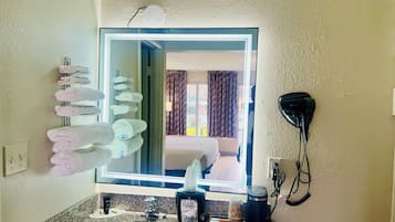A hotel bathroom with a large mirror, a towel rack with towels, a hairdryer, a sink with toiletries, and a window with curtains.