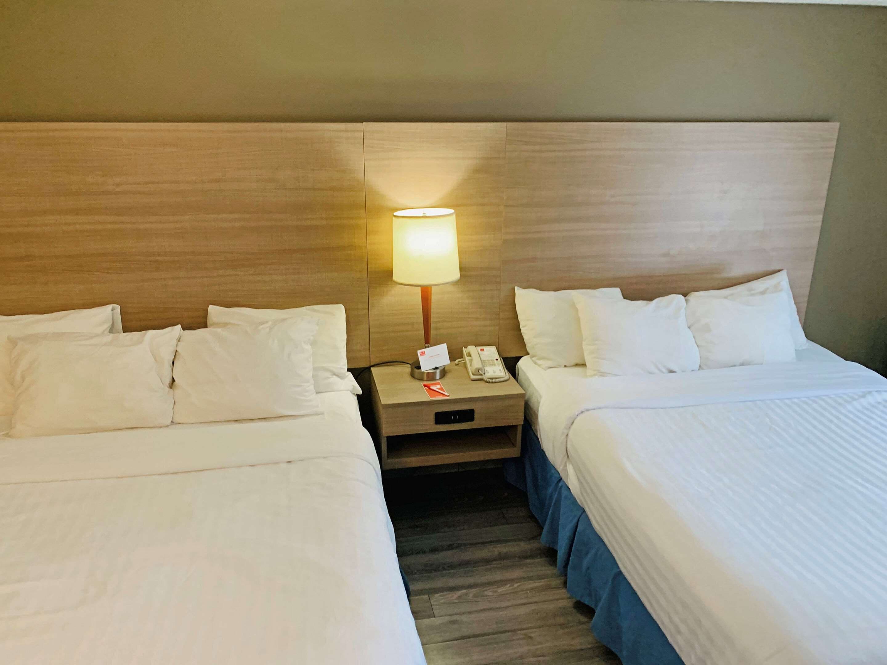 A hotel room with two beds, a nightstand with a lamp, a phone, and a card holder.