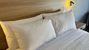Una cama bien tendida con sábanas y almohadas blancas, un cabecero de madera y una mesita de noche con una lámpara.