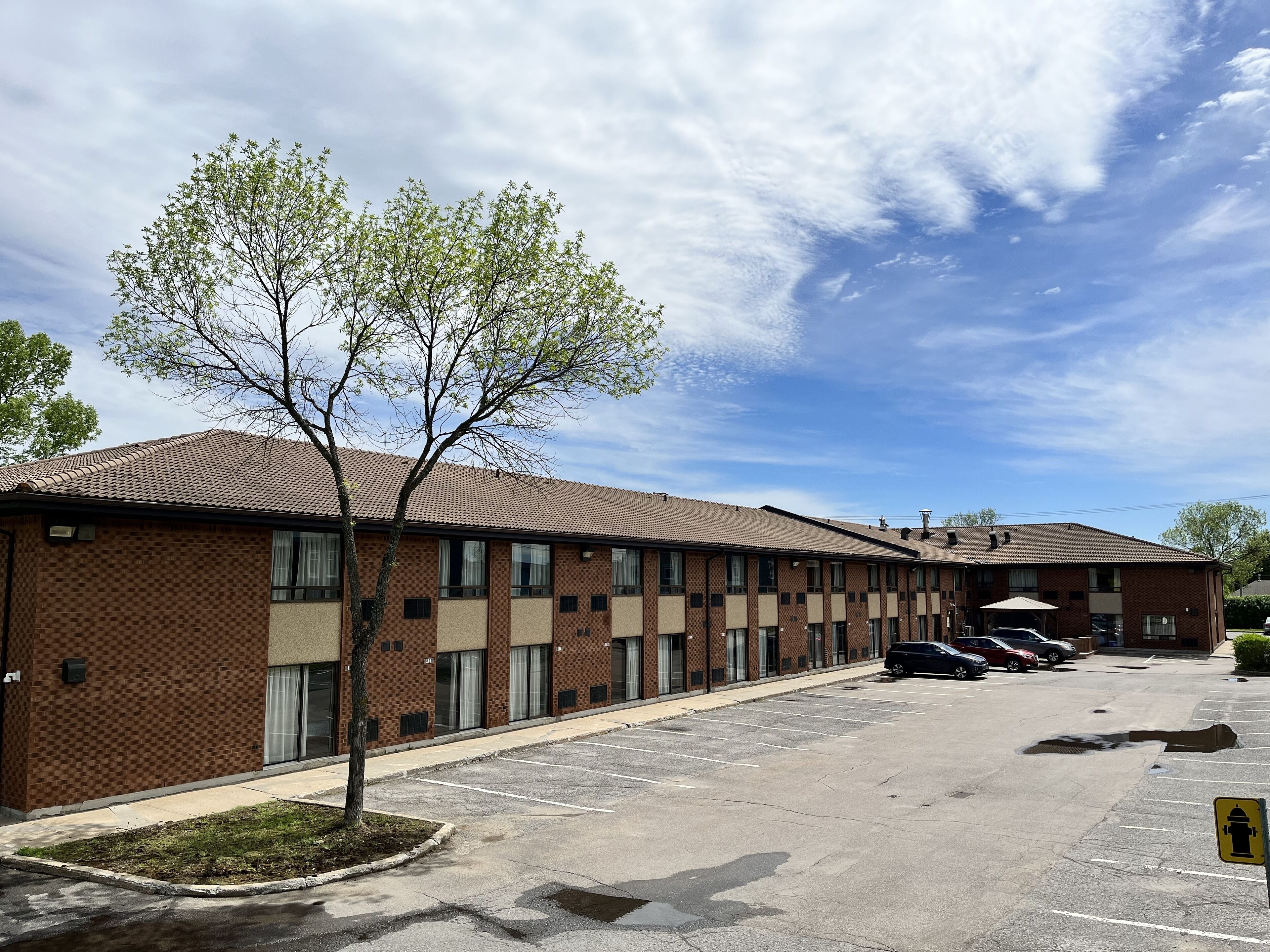 A multi-story brick building with a parking lot, a tree in front, and a clear sky.
