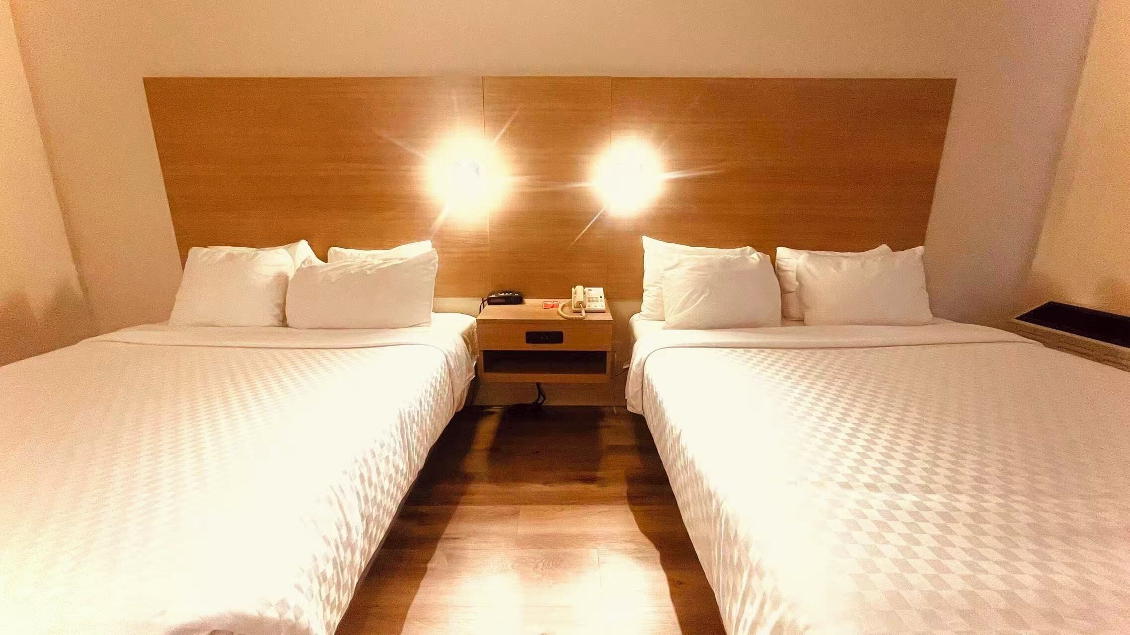 Habitación de hotel con dos camas individuales, una mesita de noche con teléfono y lámpara, y un cabecero de madera.