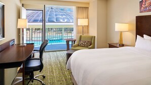 Une chambre d’hôtel avec un grand lit, un bureau, une chaise, une petite table et une vue sur une piscine, visible à travers une porte vitrée.