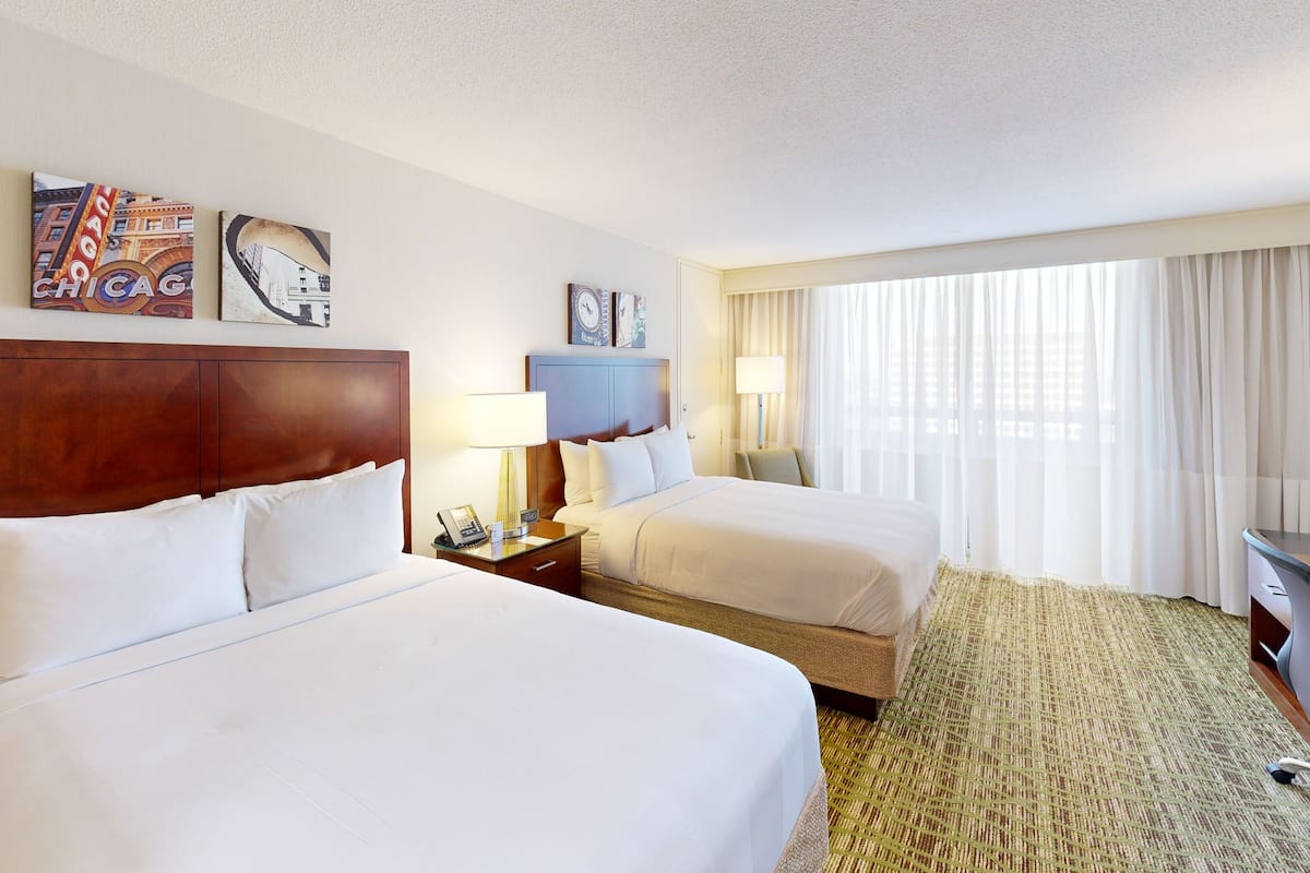 A hotel room with two beds, a desk with a chair, a telephone, and a window with curtains.