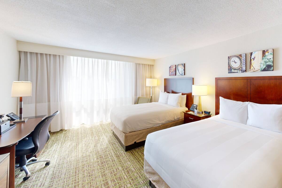 A hotel room with two beds, a desk, a chair, and a large window with curtains.