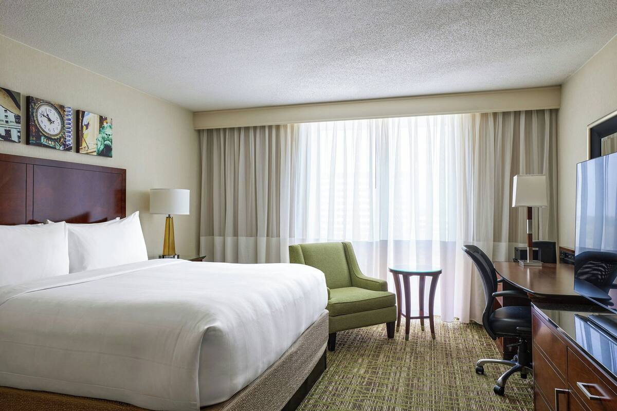 A hotel room with a large bed, a desk with a chair, a small table, and a television.