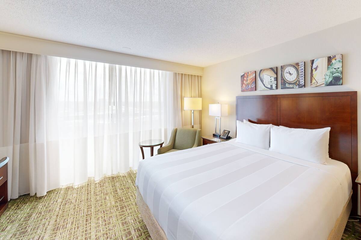 A hotel room with a large bed, a desk, a chair, a lamp, and a clock on the wall.