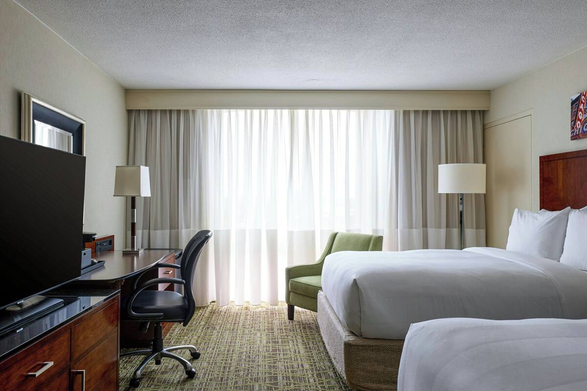 A hotel room with a bed, a desk with a TV, a chair, and a window with curtains.