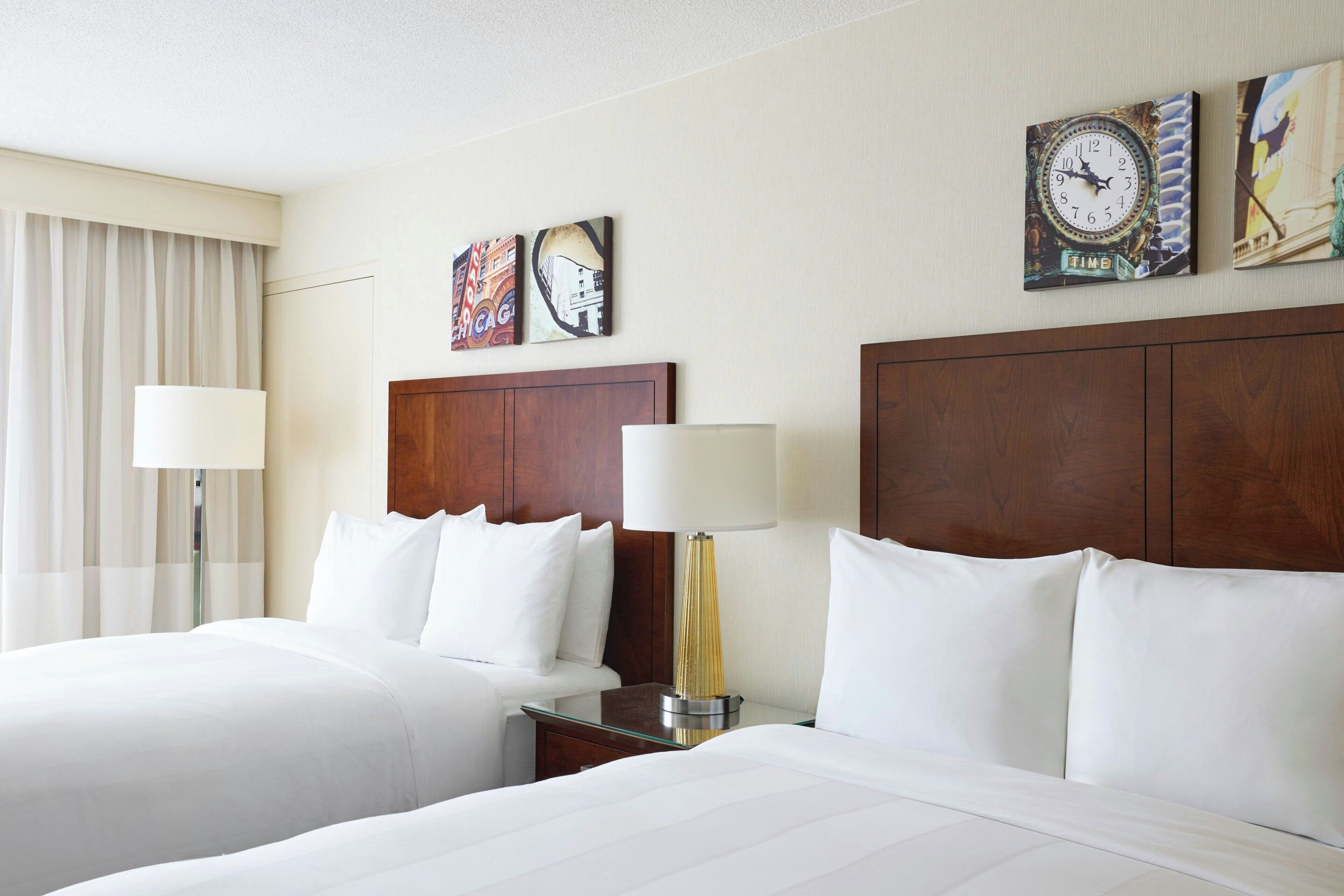 A hotel room with two beds, a wooden headboard, a nightstand with a lamp, and artwork on the wall.