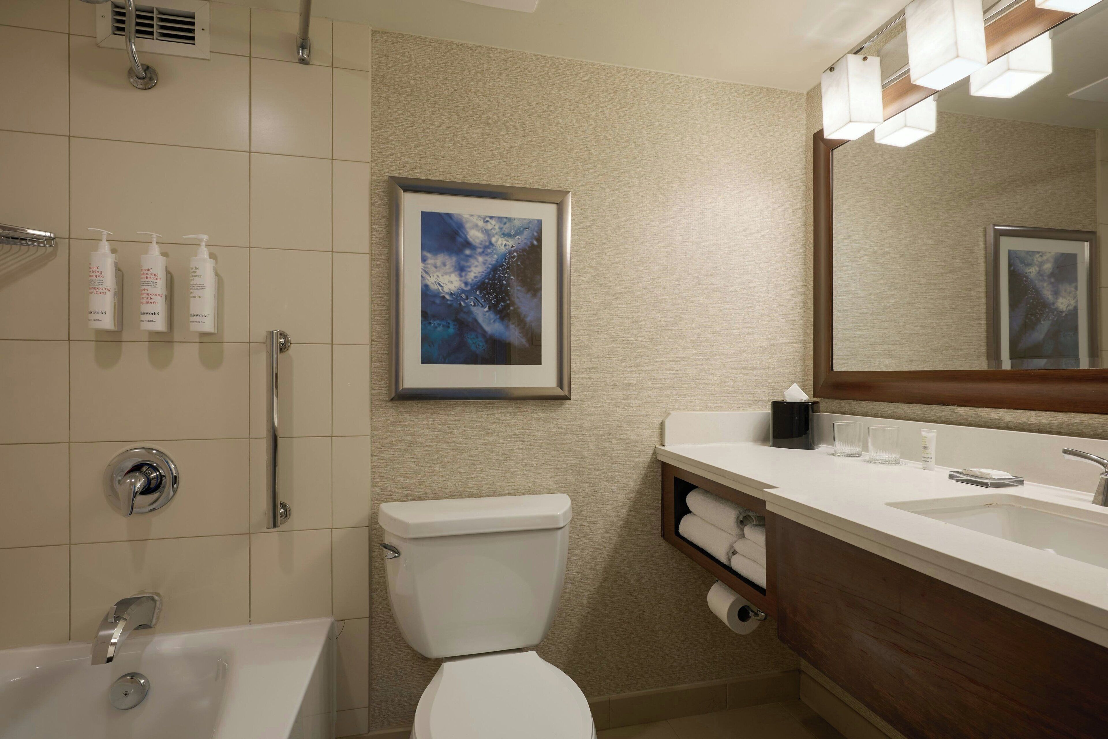 A modern bathroom with a bathtub, toilet, and sink. There are toiletries on the wall and a framed artwork above the sink.