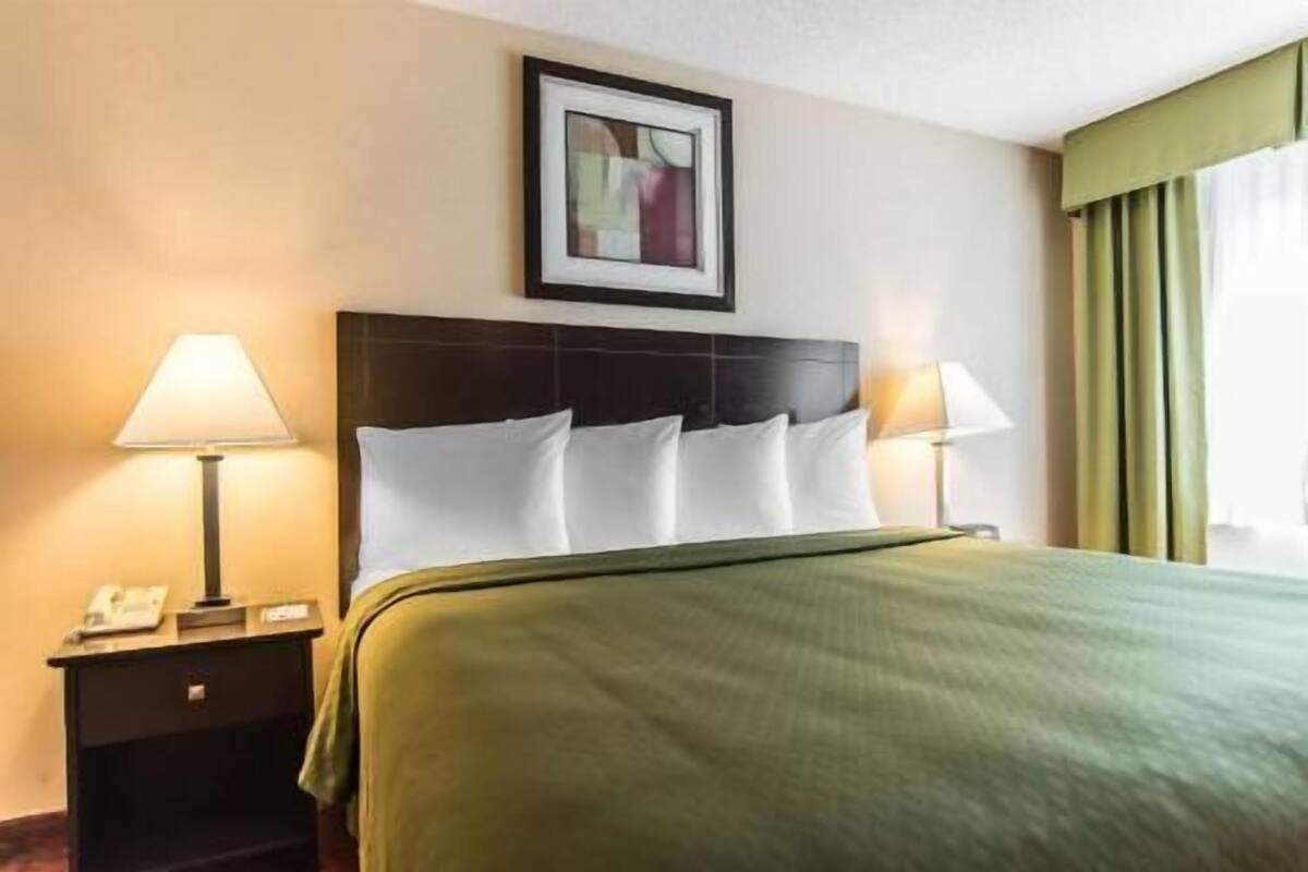 Ein Hotelzimmer mit einem Bett, zwei Nachttischlampen, einem gerahmten Bild an der Wand und einem Fenster mit Vorhängen.