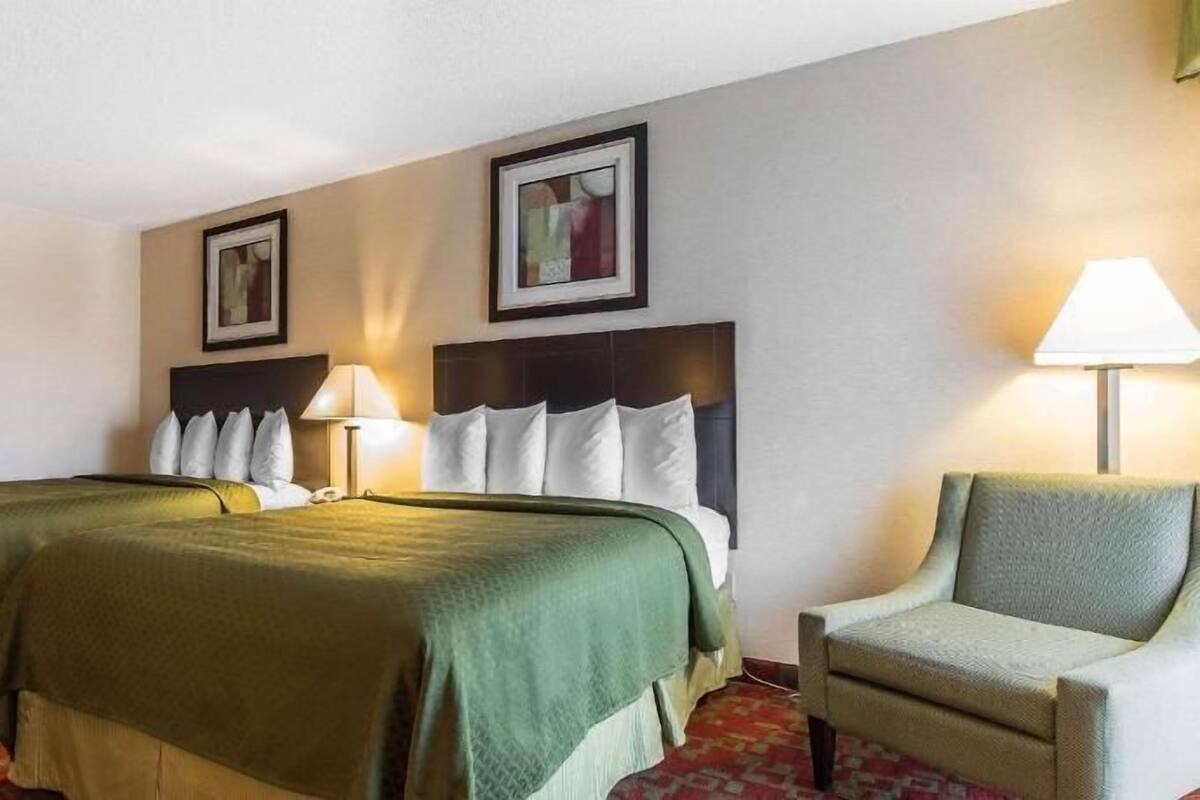 Ein Hotelzimmer mit zwei Betten, einem Sessel, einer Lampe und zwei gerahmten Bildern an der Wand.